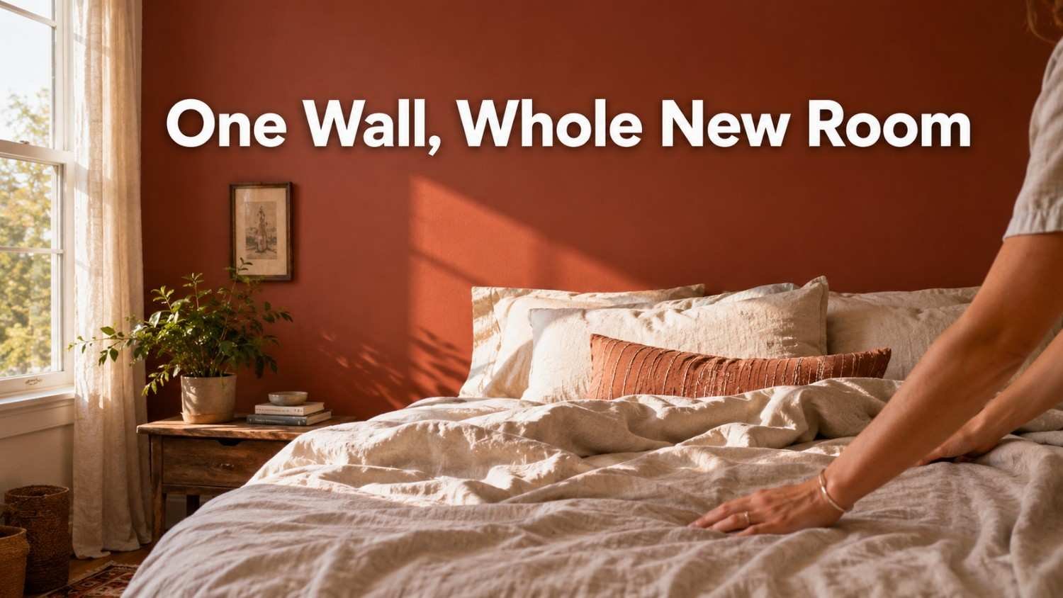 A hand smooths a linen duvet in a sunlit bedroom with a deep clay-colored accent wall, one of several inspiring bedroom decor ideas for adding warmth.