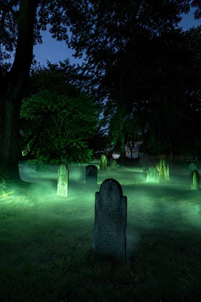Low fog covering a lawn around tombstones under green light on a calm Halloween night.