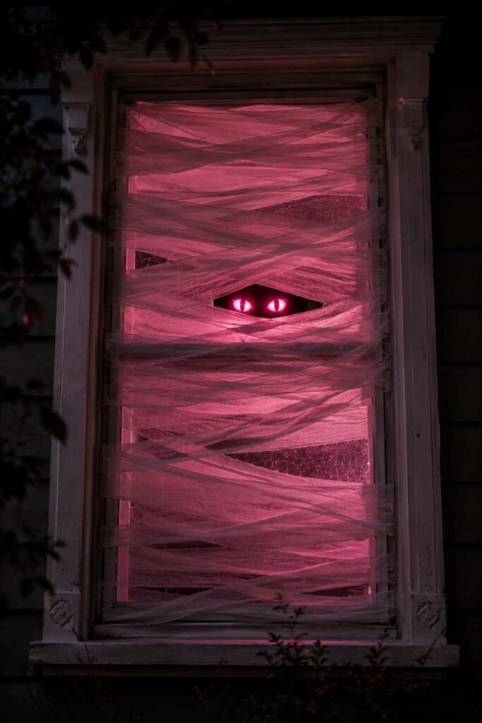 Window wrapped in white gauze with glowing pink eyes as a mummy Halloween decoration.